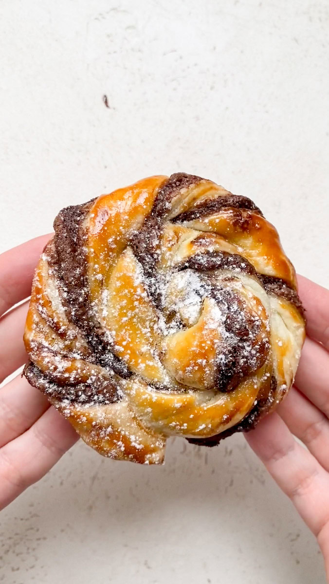 A Nutella puff pastry hold by two hand, after baking, and topped with powdered sugar.
