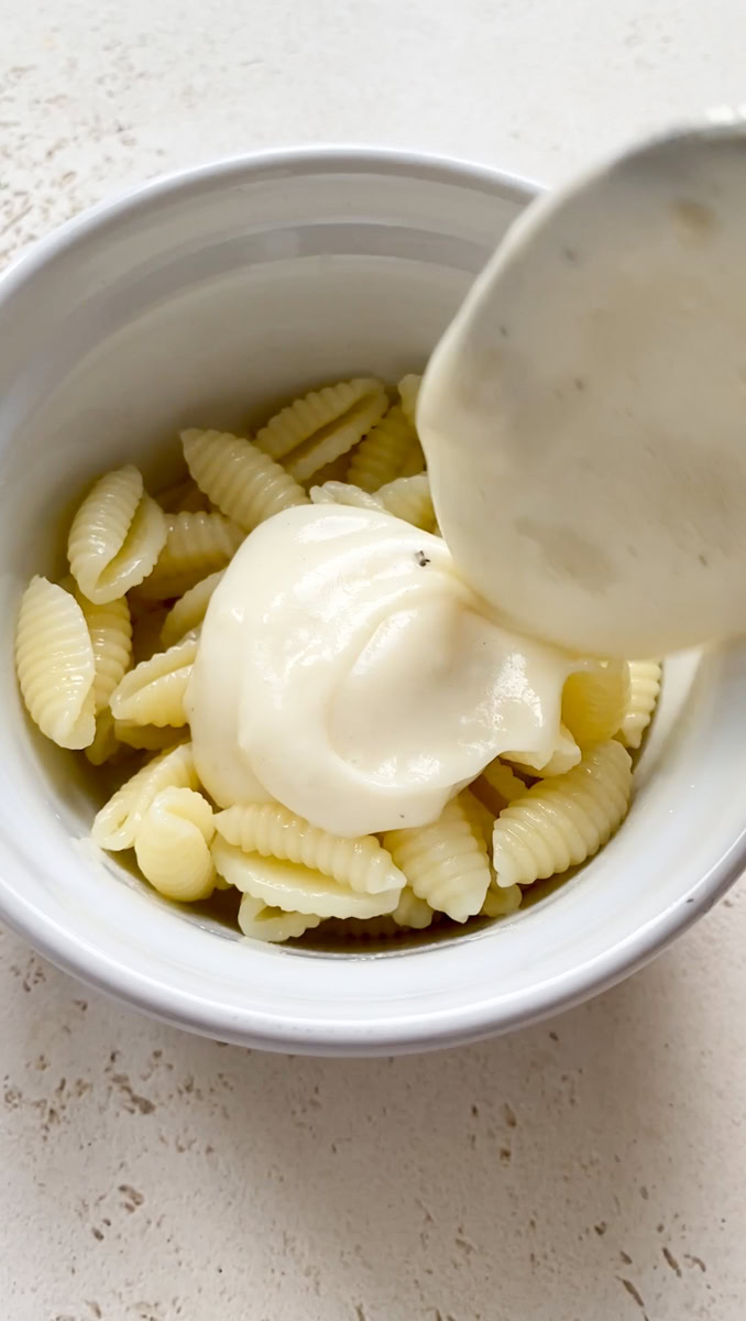 Béchamel sauce added to ramekin of pasta by a tablespoon.