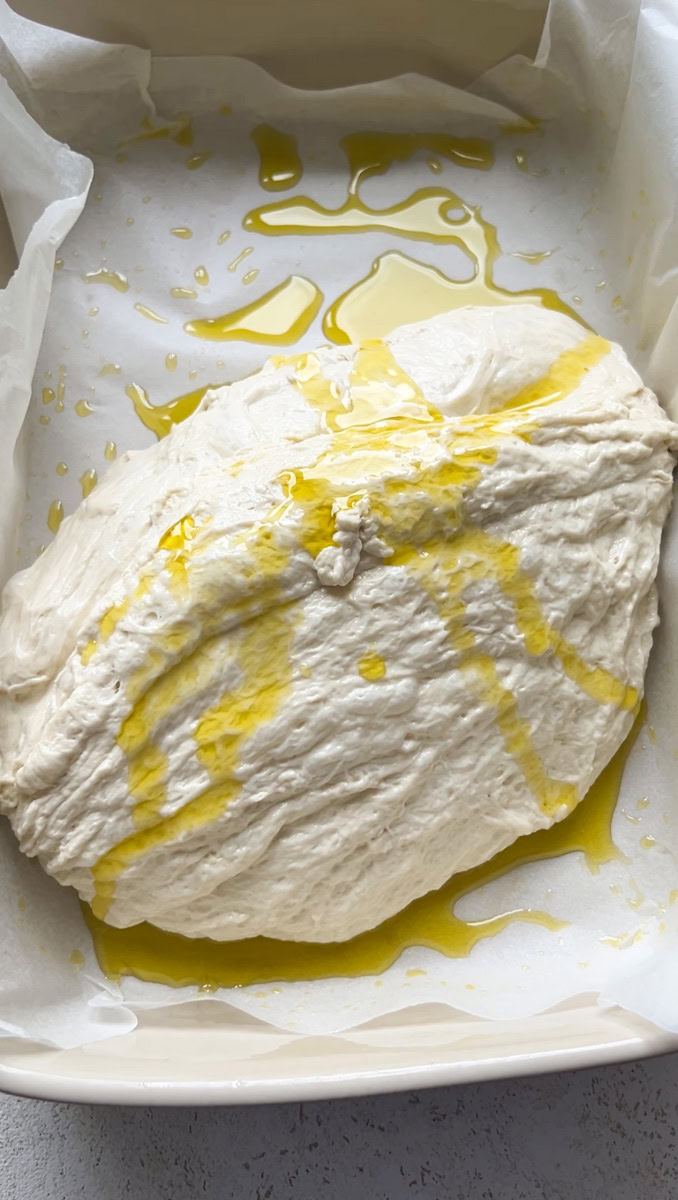 Dough topped with olive oil, in a baking dish with parchment paper and olive oil.