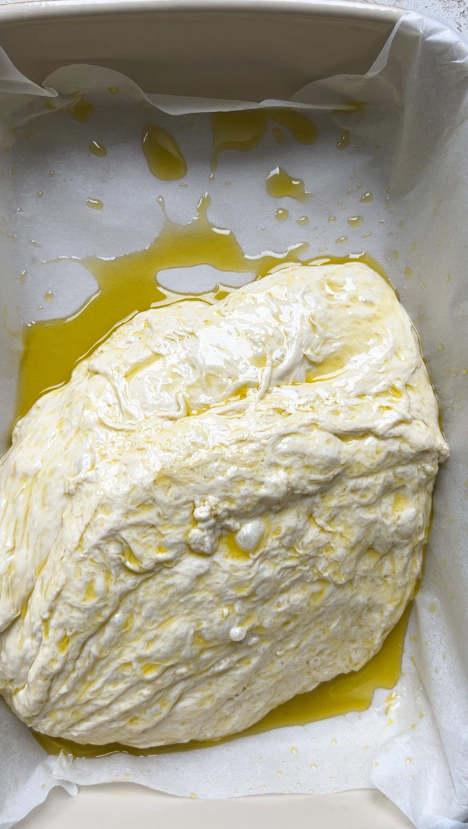Dough, topped with olive oil, in a baking dish with parchment paper and olive oil.
