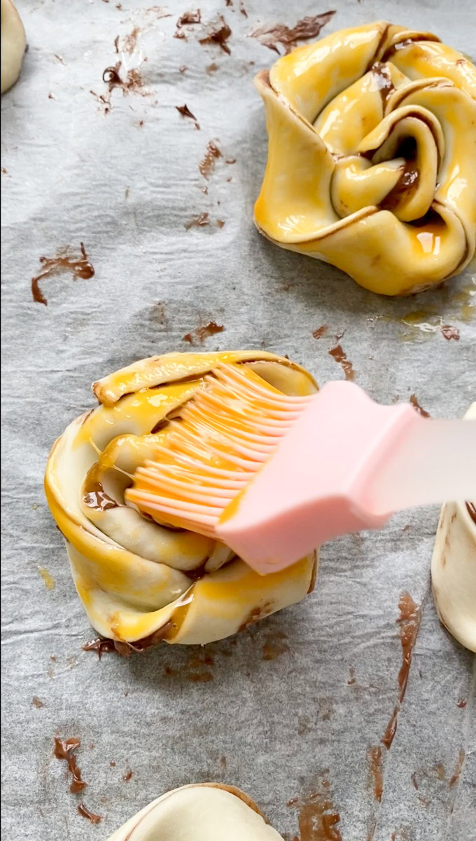 A pink silicone pastry brush brushing beaten egg yolk on a twisted puff pastry.