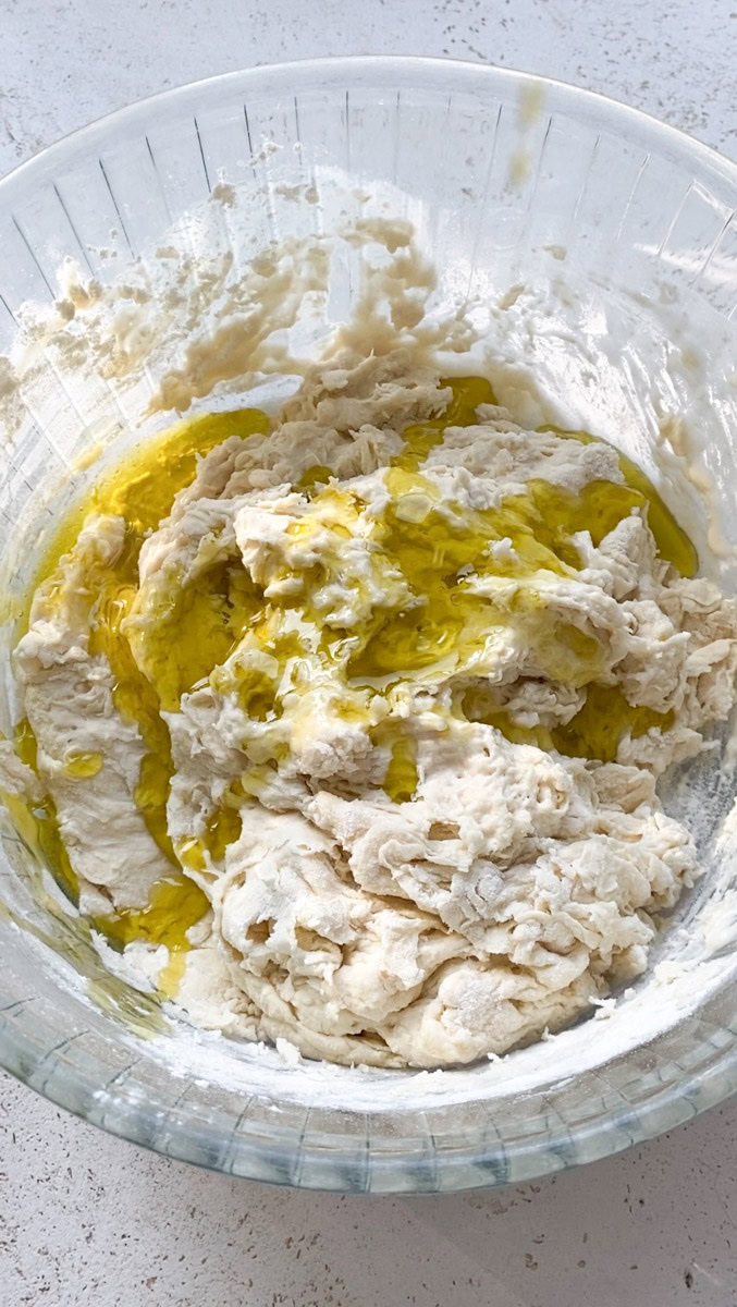 Olive oil added to the dough in the large transparent bowl.