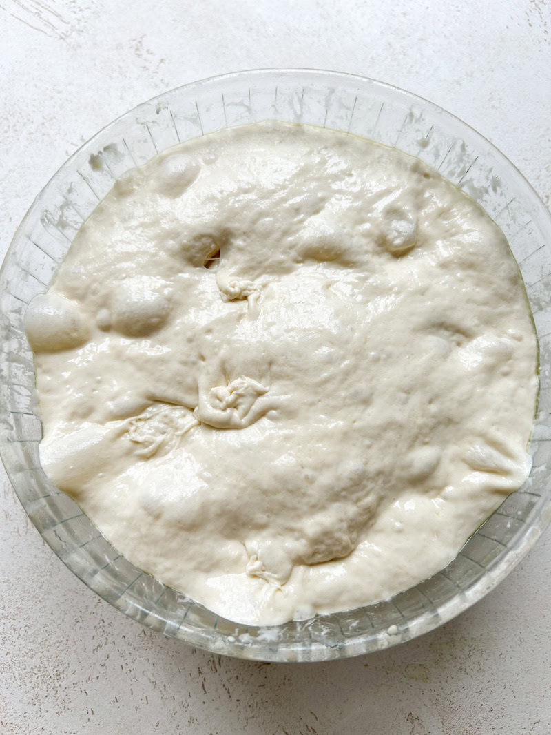 Focaccia dough with air bubbles after its first rise, in the large transparent bowl.