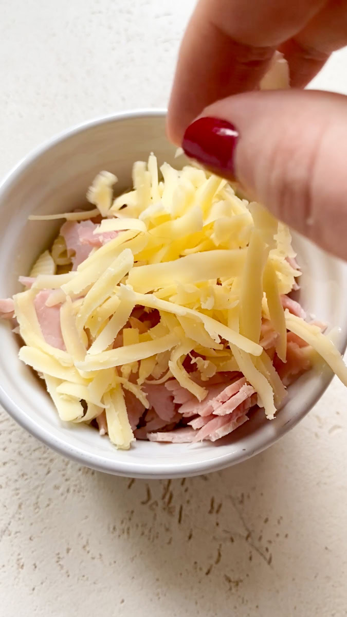 A hand that adds grated cheese to the ramekin of pasta, béchamel and ham.