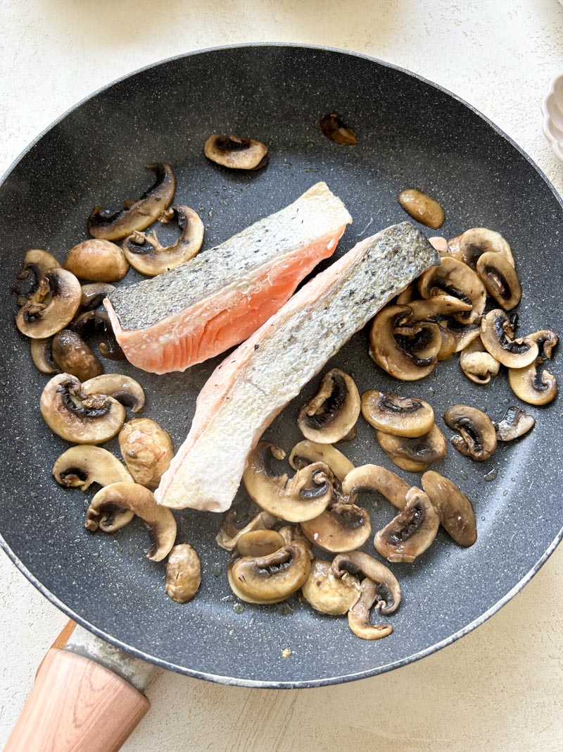 Sliced cremini mushrooms with two salmon fillets in a large grey pan.