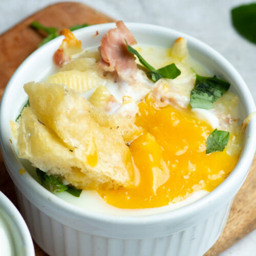 Egg cocotte in a white ramekin with a piece of focaccia that drips with egg yolk.