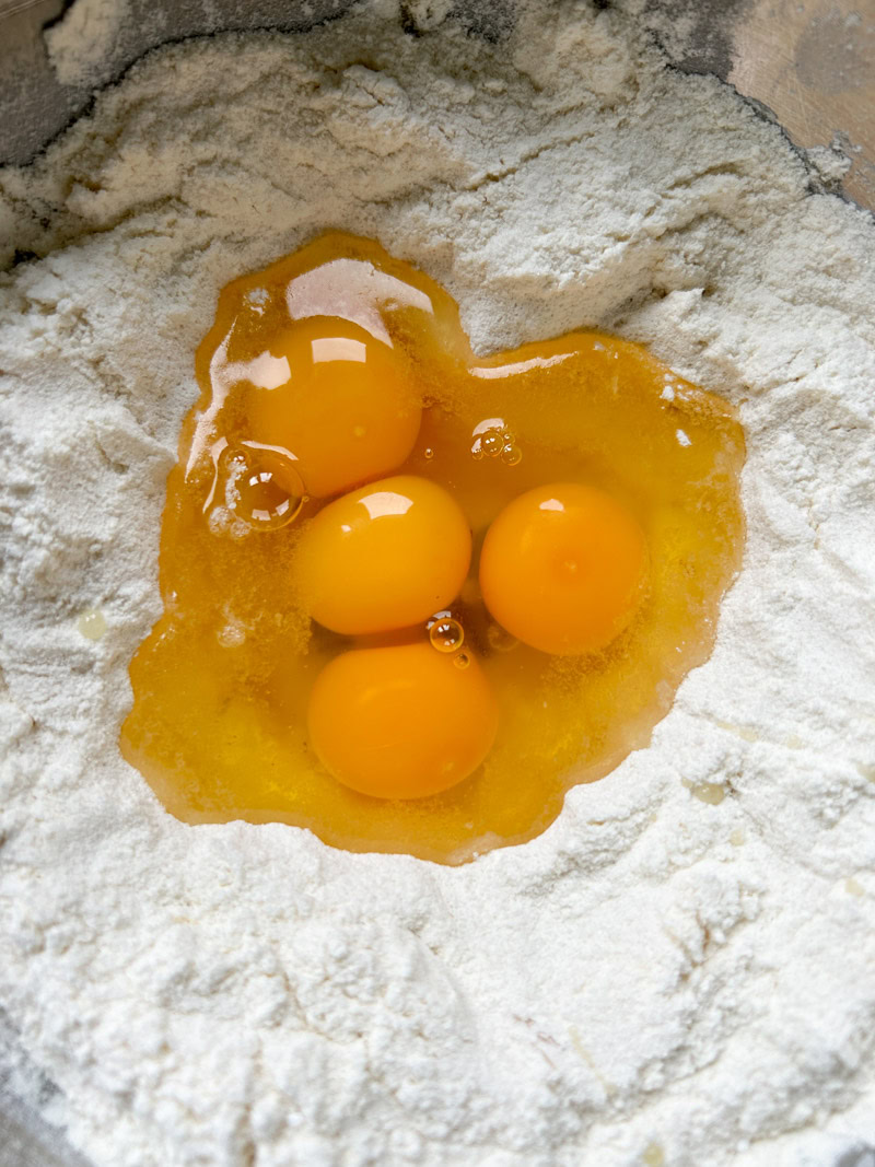 Four eggs are added to the flour well in the stainless steel bowl.