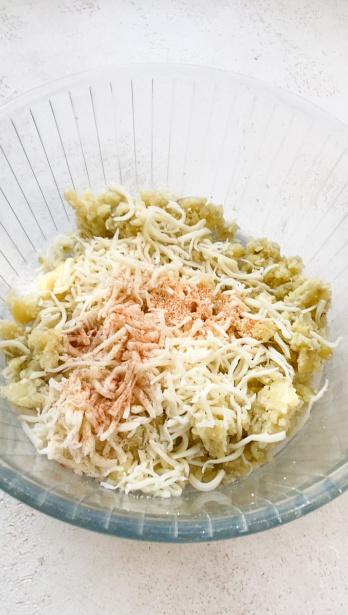 Shredded cheese and spices are added to the mashed potatoes in the transparent bowl.