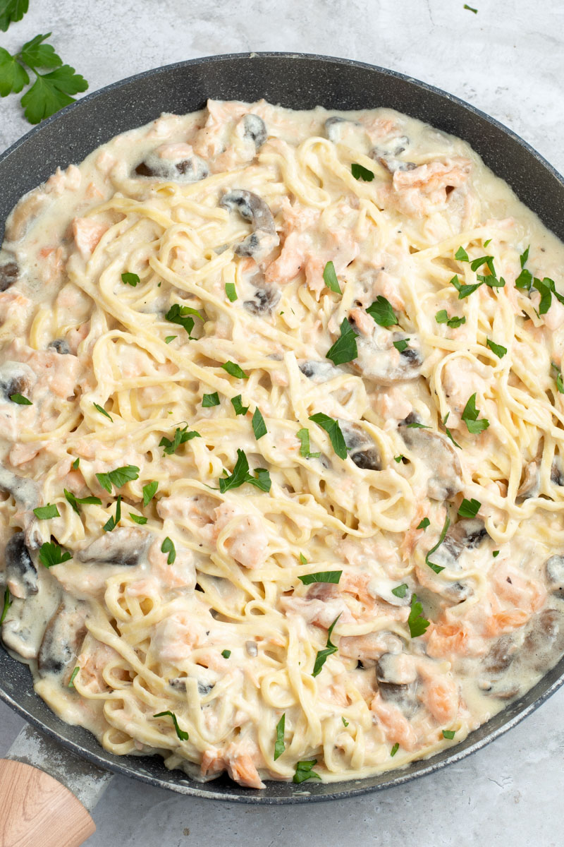 Salmon Alfredo pasta in a large skillet with fresh parsley and cremini mushrooms.