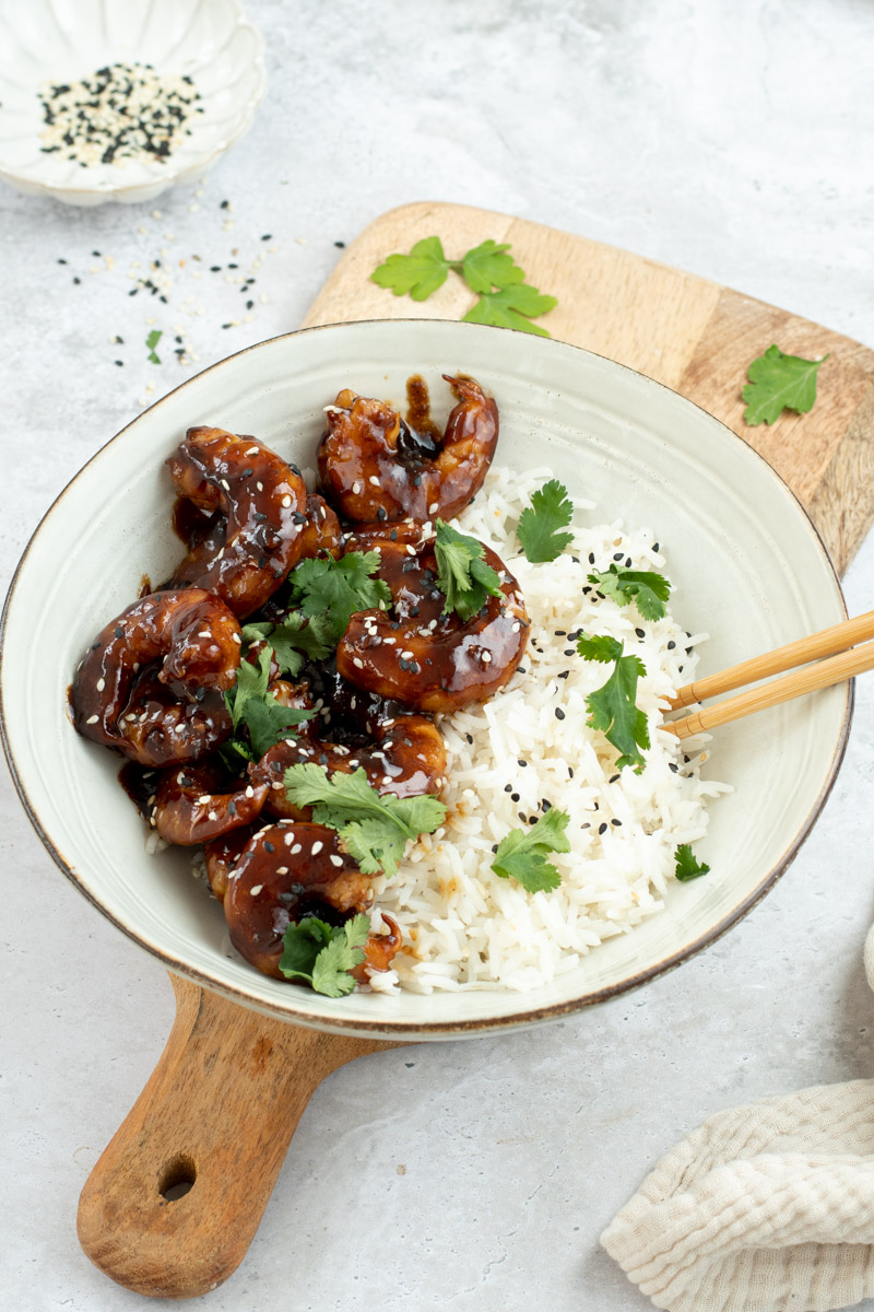 Teriyaki shrimp in a beige bowl with rice, sesame seeds, fresh cilantro and wooden chopsticks.