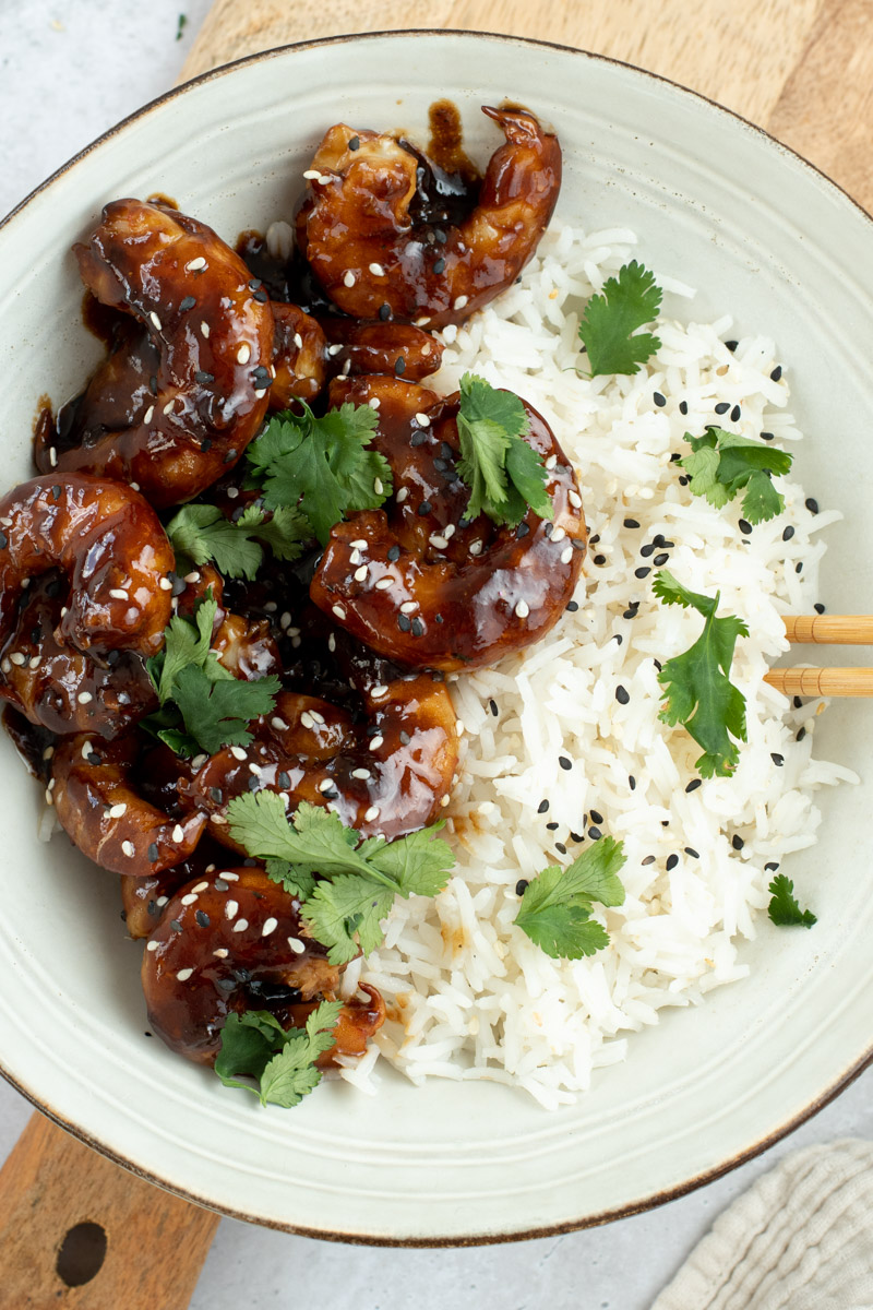 Teriyaki shrimp in a beige bowl with rice, sesame seeds, fresh cilantro and wooden chopsticks.