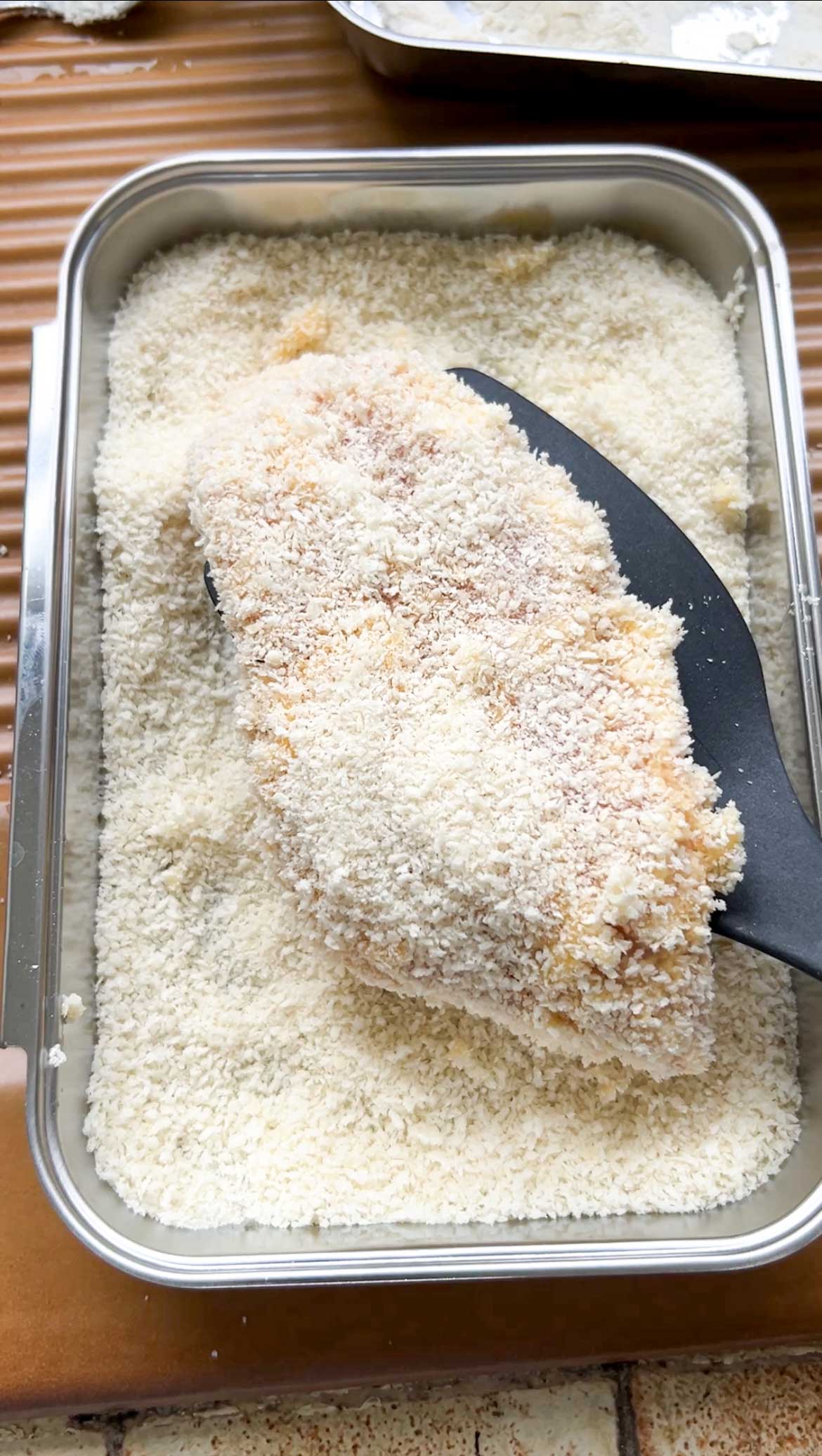 Chicken fillet held by tongs and dipped in a dish of Panko breadcrumbs.