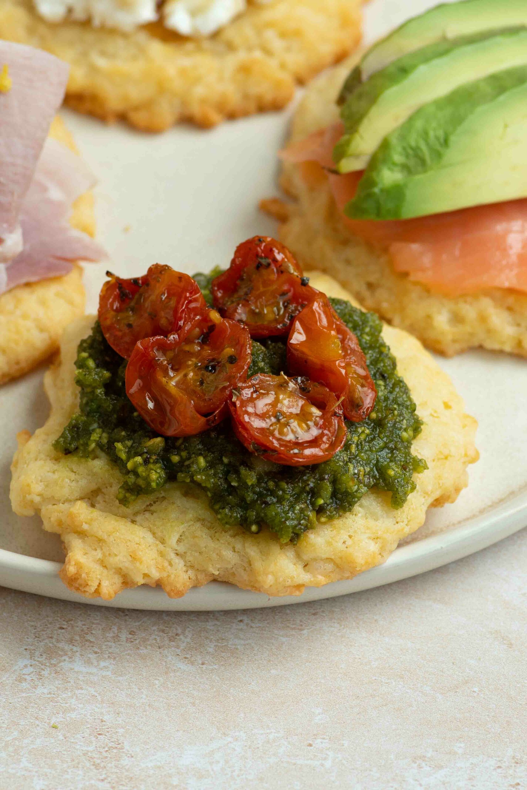 A cookie on a plate, topped with pesto and confit cherry tomato halves.
