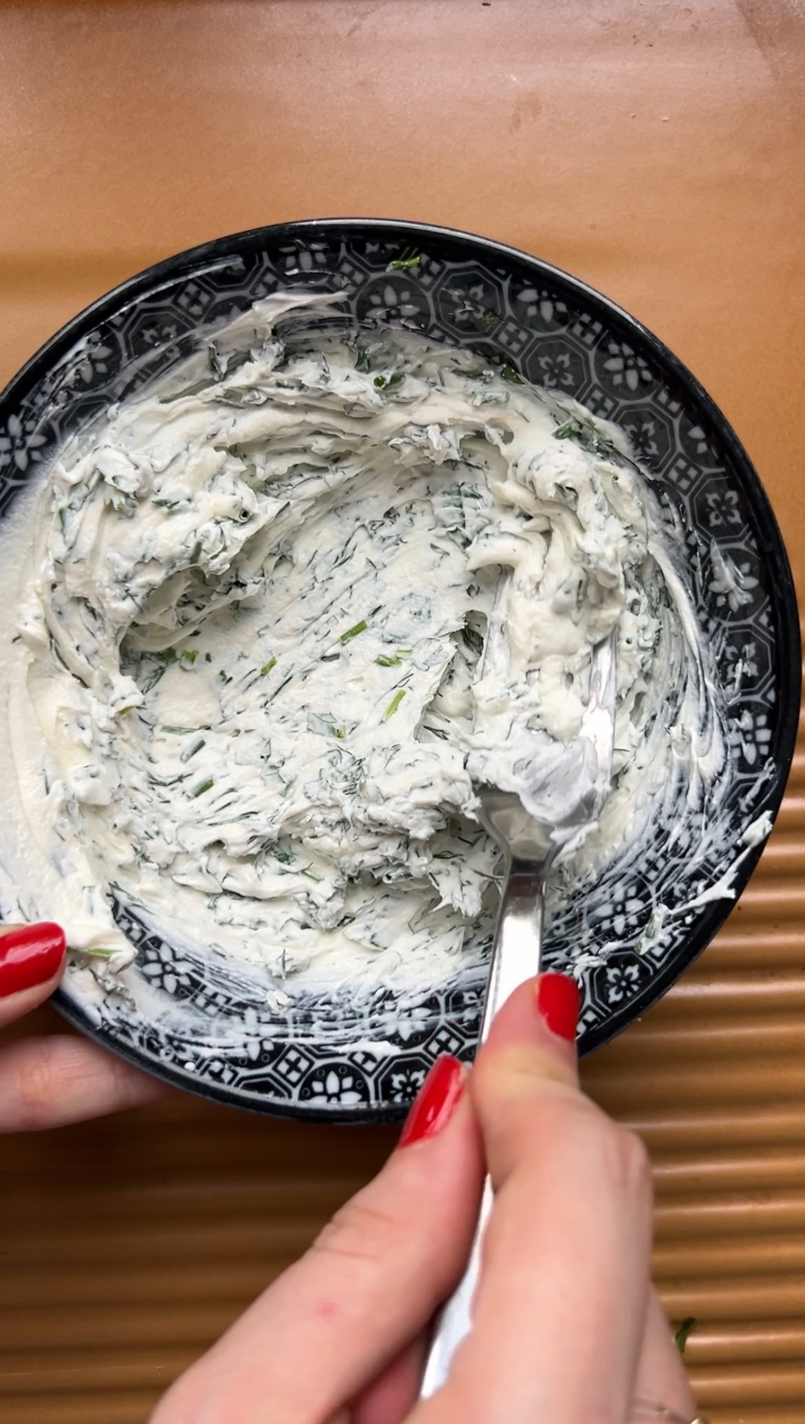A hand mixing cream cheese and chopped herbs in a bowl, with a fork.