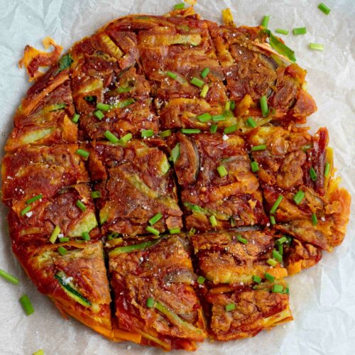 A large Korean pancake placed on baking paper, cut into equal-sized squares and sprinkled with fresh chives.