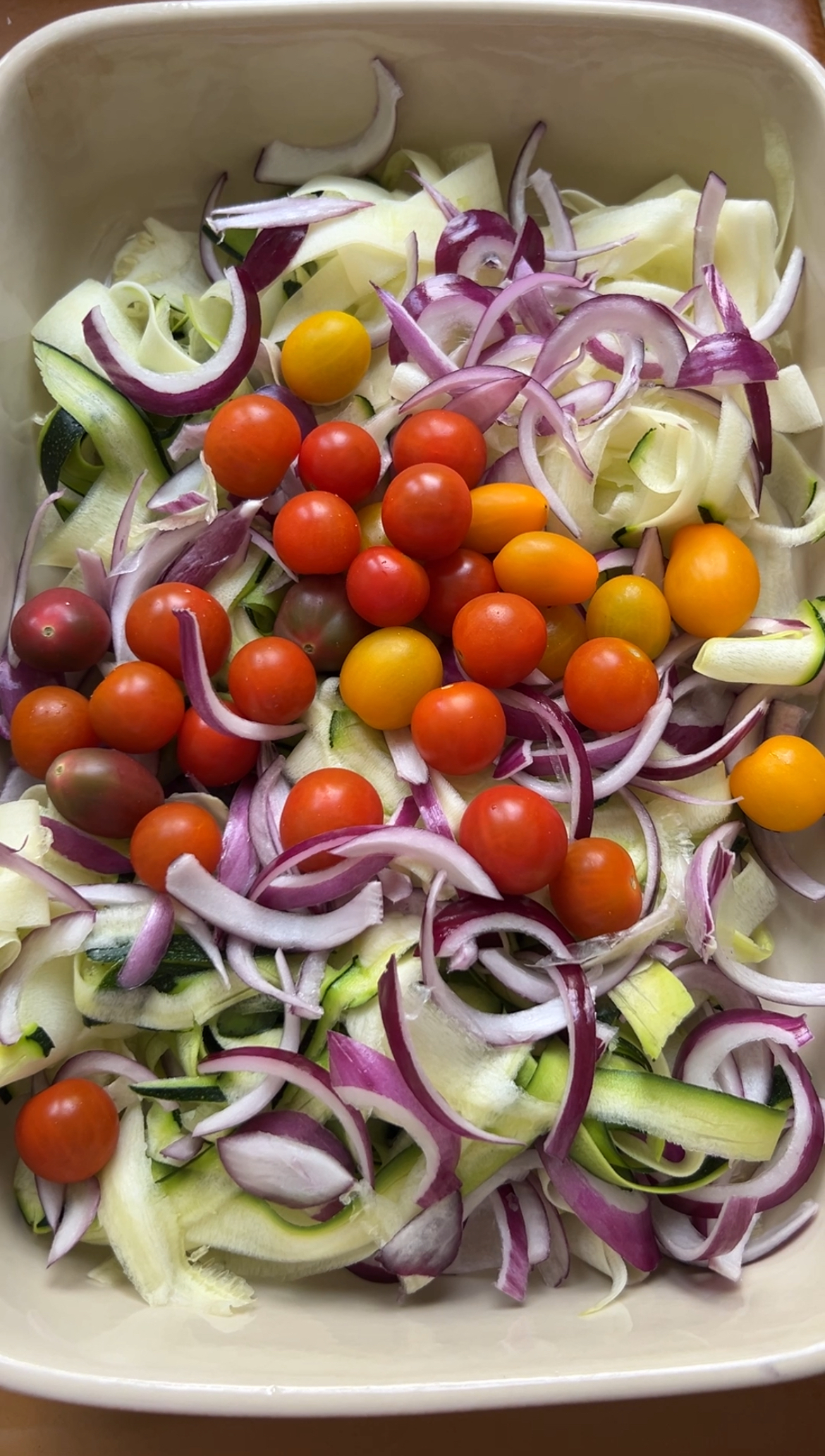 Zucchini tagliatelle, red onion strips and cherry tomatoes are in a large ovenproof dish.