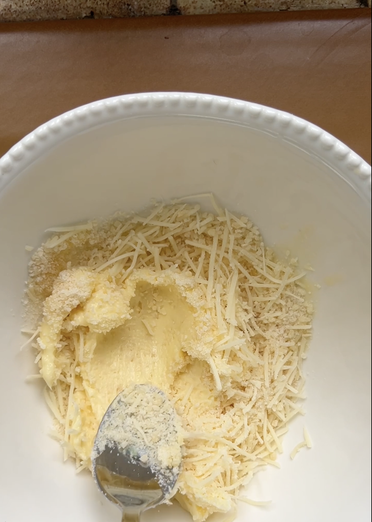 Beurre mou, Parmesan et gruyère râpés dans un grand bol blanc, mélangés par une cuillère à soupe.