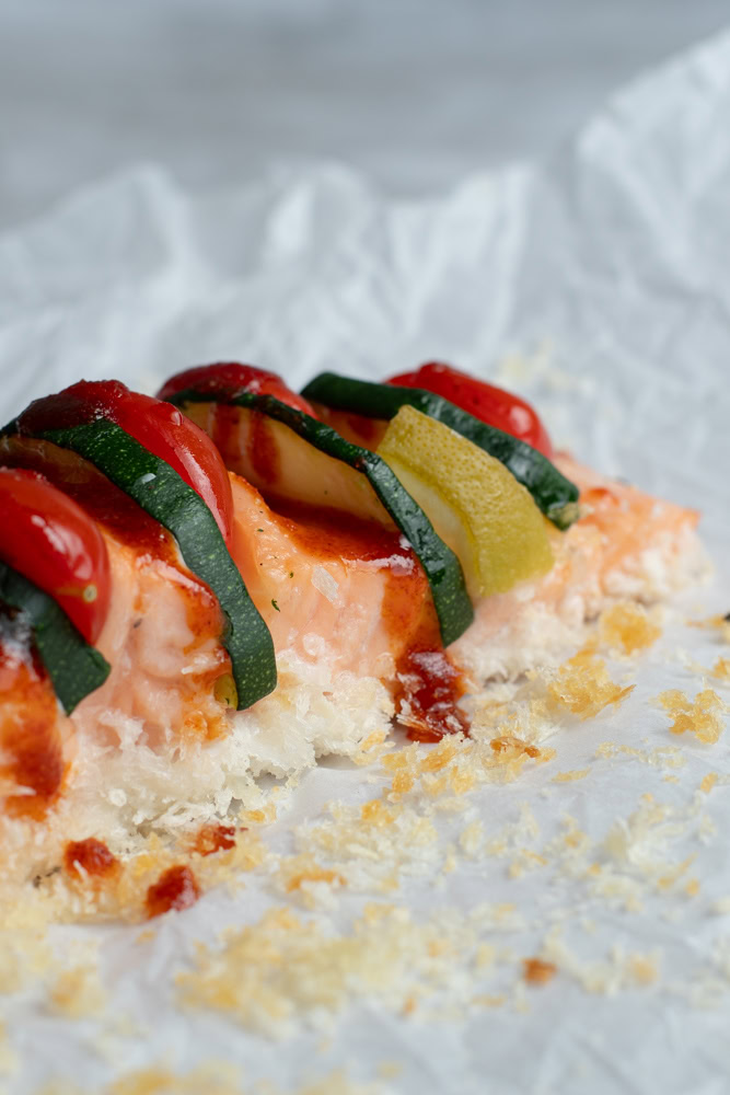 Hasselback salmon garnished with cherry tomatoes, zucchini strips and lime wedges on a sheet of parchment paper.