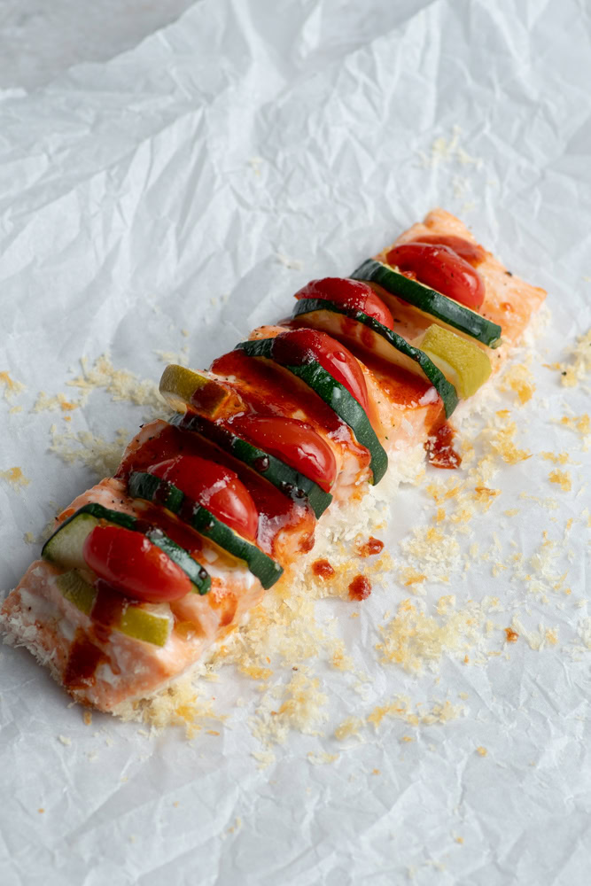 Hasselback salmon garnished with cherry tomatoes, zucchini strips and lime wedges on a sheet of parchment paper.