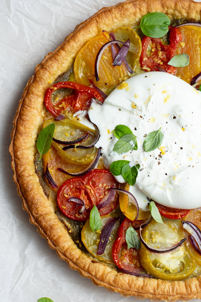 Multicolored tomato tart on parchment paper, with a large burrata in the middle and lots of fresh basil leaves.