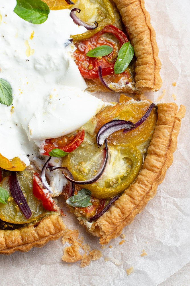 Multicolored slice of tomato tart on parchment paper, with a large burrata in the middle and lots of fresh basil leaves.