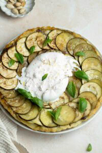 Zucchini tatin on a plate with burrata, mint and basil leaves and a small plate of pistachios at top left.