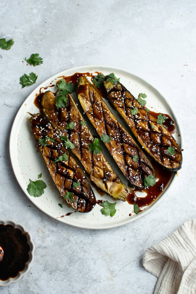 Four zucchini halves on a white plate with Teriyaki sauce, sesame seeds and fresh cilantro.