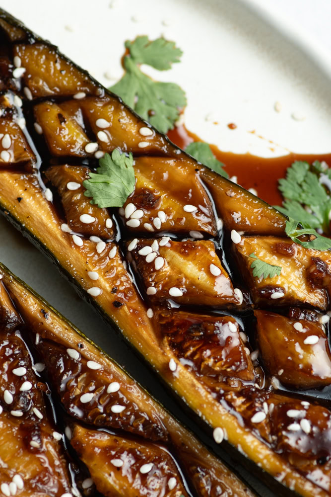 Zucchini halves in a white plate with Teriyaki sauce, sesame seeds and fresh cilantro leaves.