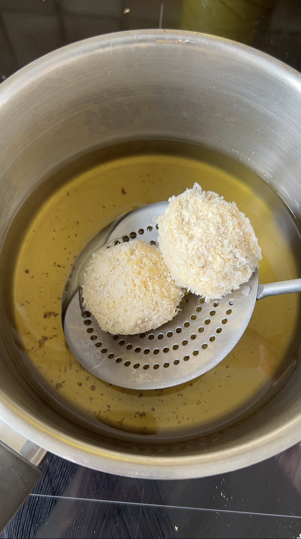 Two croquetas, held by a skimmer, are plunged into a pan of frying oil.