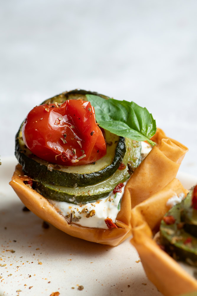 Cup croustillante dans une assiette beige garnie de fromage frais et légumes confits (courgettes, tomates cerises et échalotes) avec une feuille de basilic.