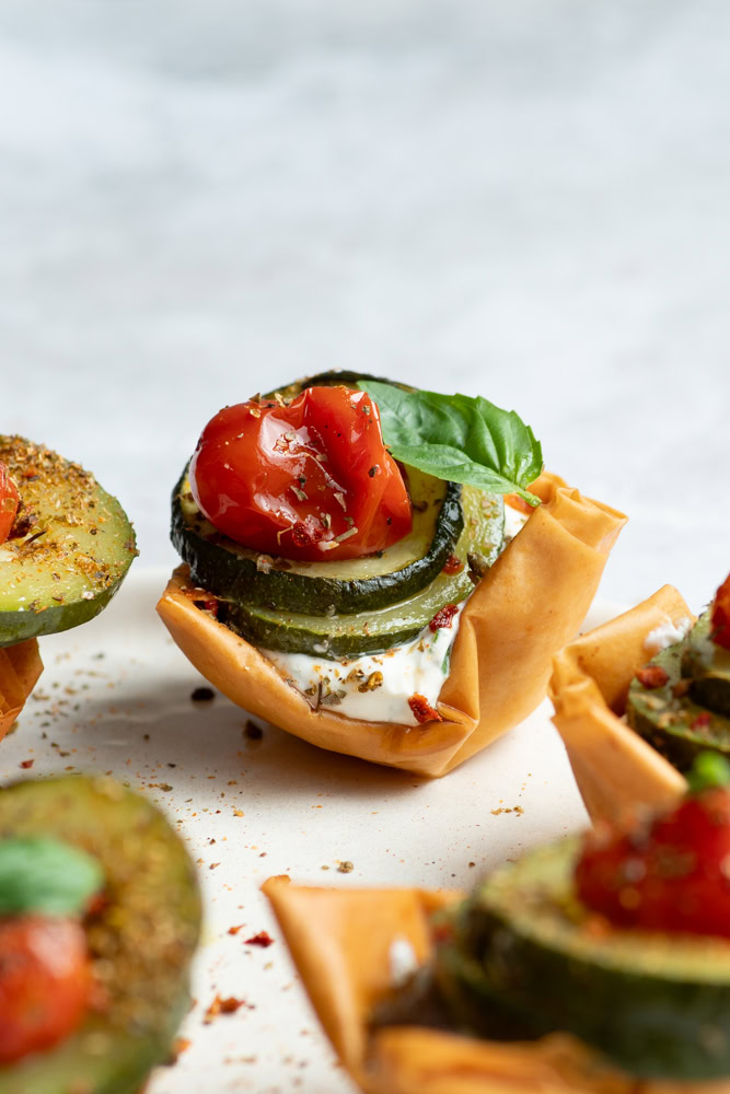 Cups croustillantes dans une assiette beige garnies de fromage frais et légumes confits (courgettes, tomates cerises et échalotes) avec une feuille de basilic.