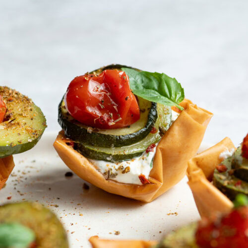 Crispy cups in a beige plate garnished with cream cheese and confit vegetables (zucchini, cherry tomatoes and shallots) with a basil leaf.