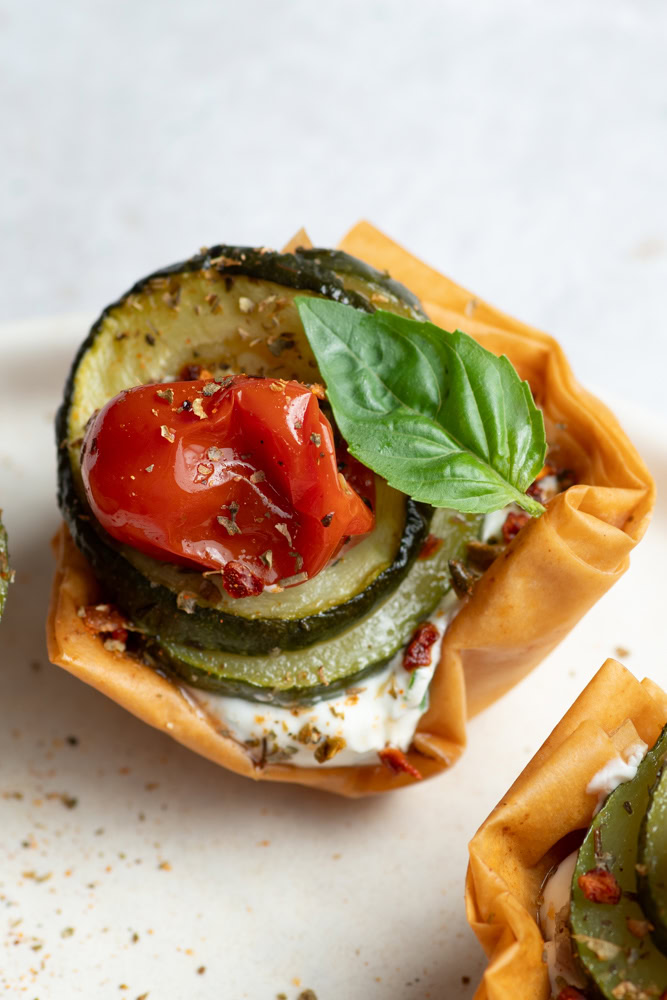 Cup croustillante dans une assiette beige garnie de fromage frais et légumes confits (courgettes, tomates cerises et échalotes) avec une feuille de basilic.