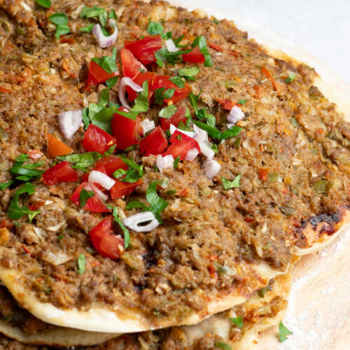 Several Lahmacun on a wooden board with chopped fresh parsley, chunks of tomato and a bowl of paprika.