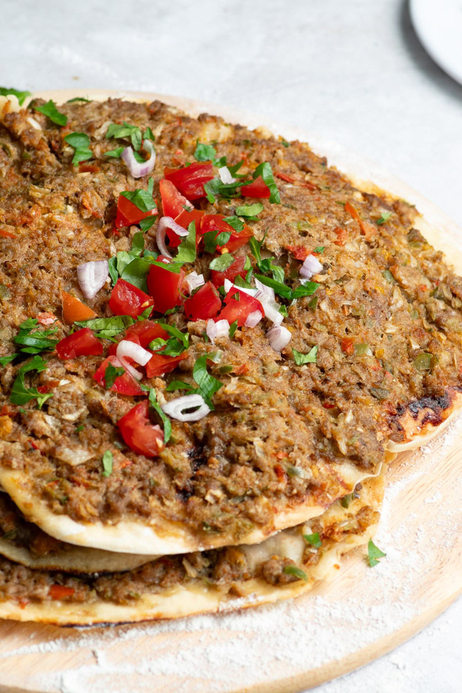 Several Lahmacun on a wooden board with chopped fresh parsley, chunks of tomato and a bowl of paprika.