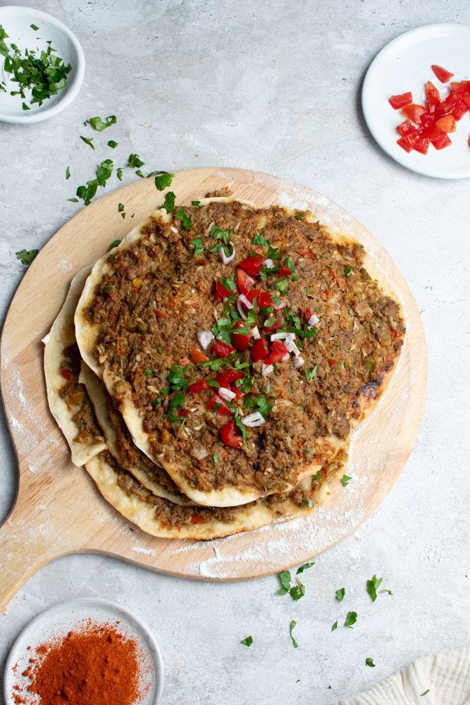 Several Lahmacun on a wooden board with chopped fresh parsley, chunks of tomato and a bowl of paprika.