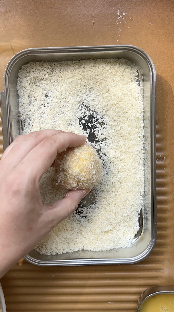 The ball of Mac and Cheese is held in one hand and dipped in Panko breadcrumbs.