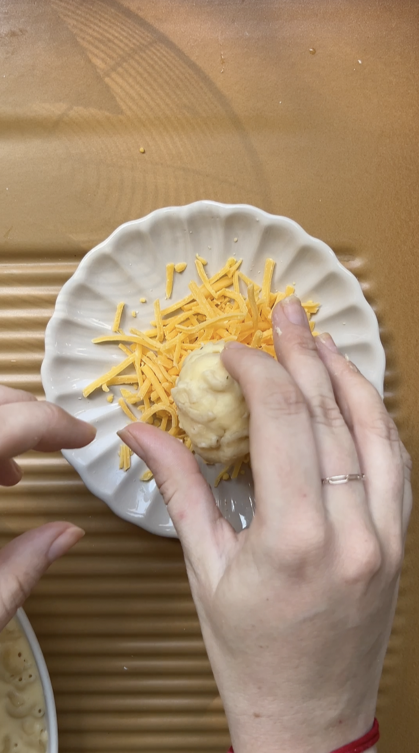 Deux mains passent une boule de Mac and cheese dans une petite assiette de cheddar râpé.