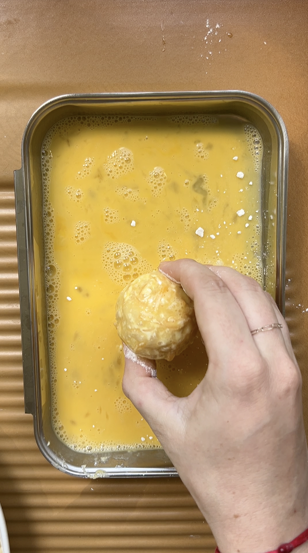 La boule de Mac and cheese est tenue par une main et passée dans les oeufs battus.