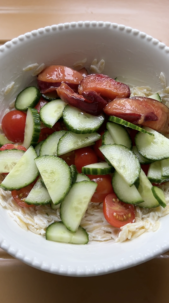 Lamelles de courgette et de concombre, tomates cerises et quartiers de pêche rajoutés aux pâtes dans le grand saladier blanc.