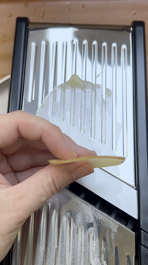 A very thin slice of potato held by one hand over a mandolin.