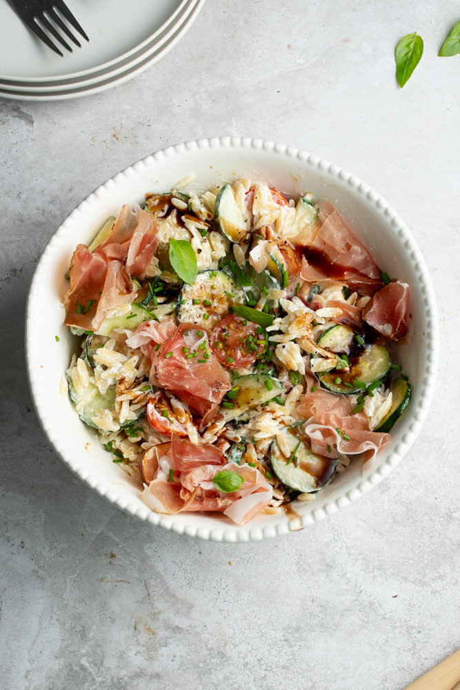 Salade de pâtes dans un grand saladier blanc, avec plein de légumes et du jambon de Parme.