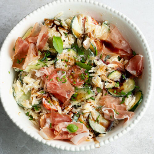 Salade de pâtes dans un grand saladier blanc, avec plein de légumes et du jambon de Parme.