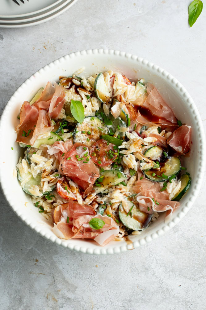 Pasta salad in a large white salad bowl, with lots of vegetables and Parma ham.