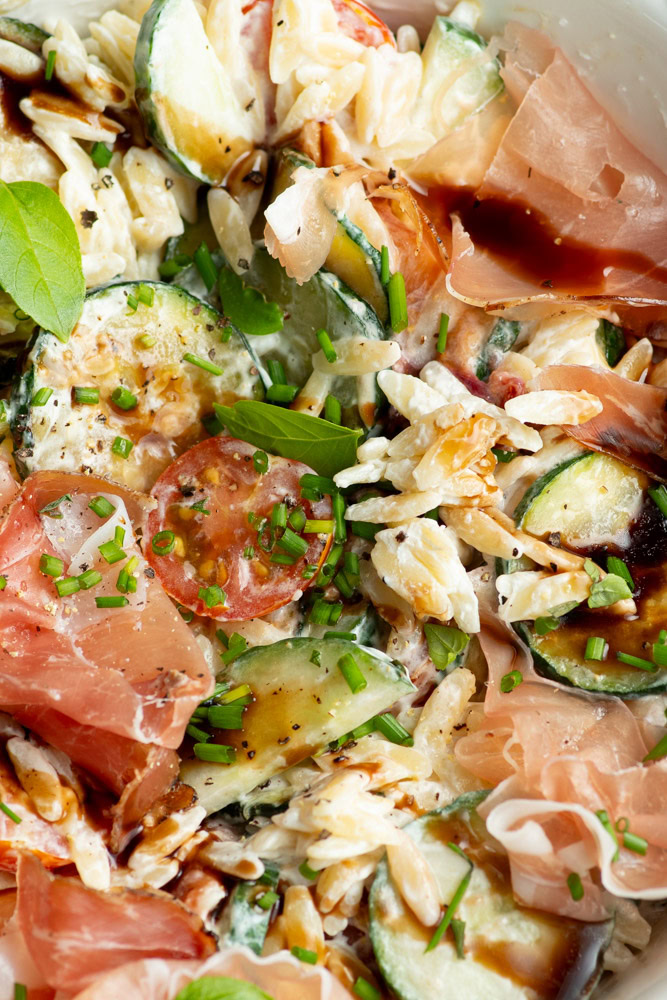 Pasta salad in a large white salad bowl, with lots of vegetables and Parma ham.