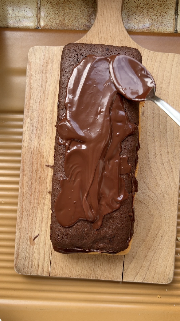 Génoise au chocolat sur la première génoise nature. Une main étale la ganache au chocolat dessus.