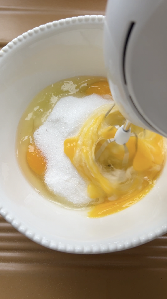 Eggs and sugar in a white bowl, mixed with an electric mixer.